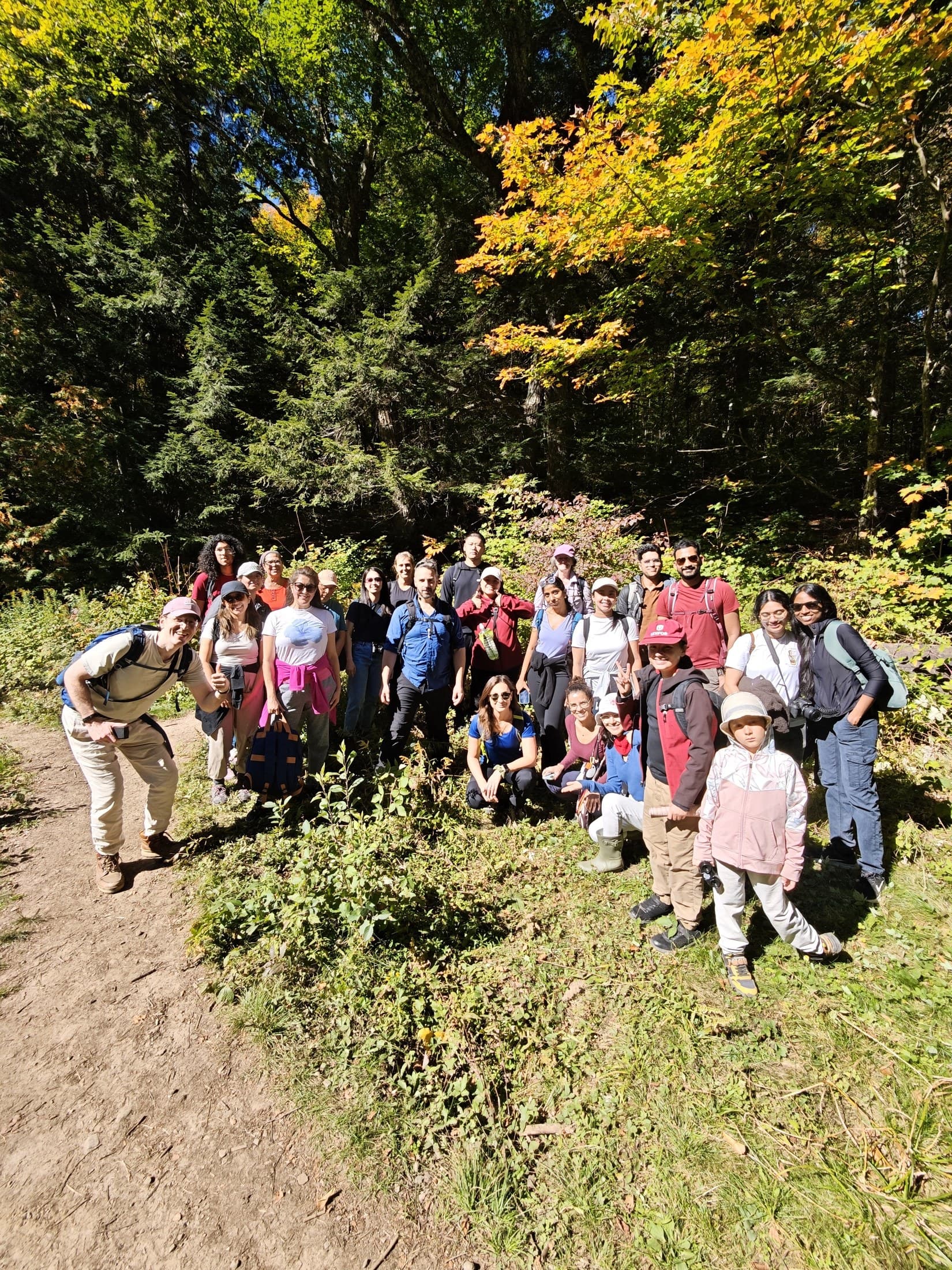 About twenty five people posing together on a trail in Algonquin in front of yellow and green maples, waving at the camera