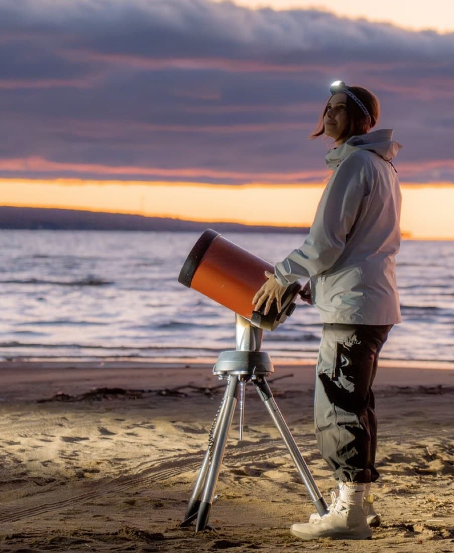 Astronomer Caroline El-Khoury in a headlamp beside her telescope on a beach, looking up at an orange and purple sky just after sunset