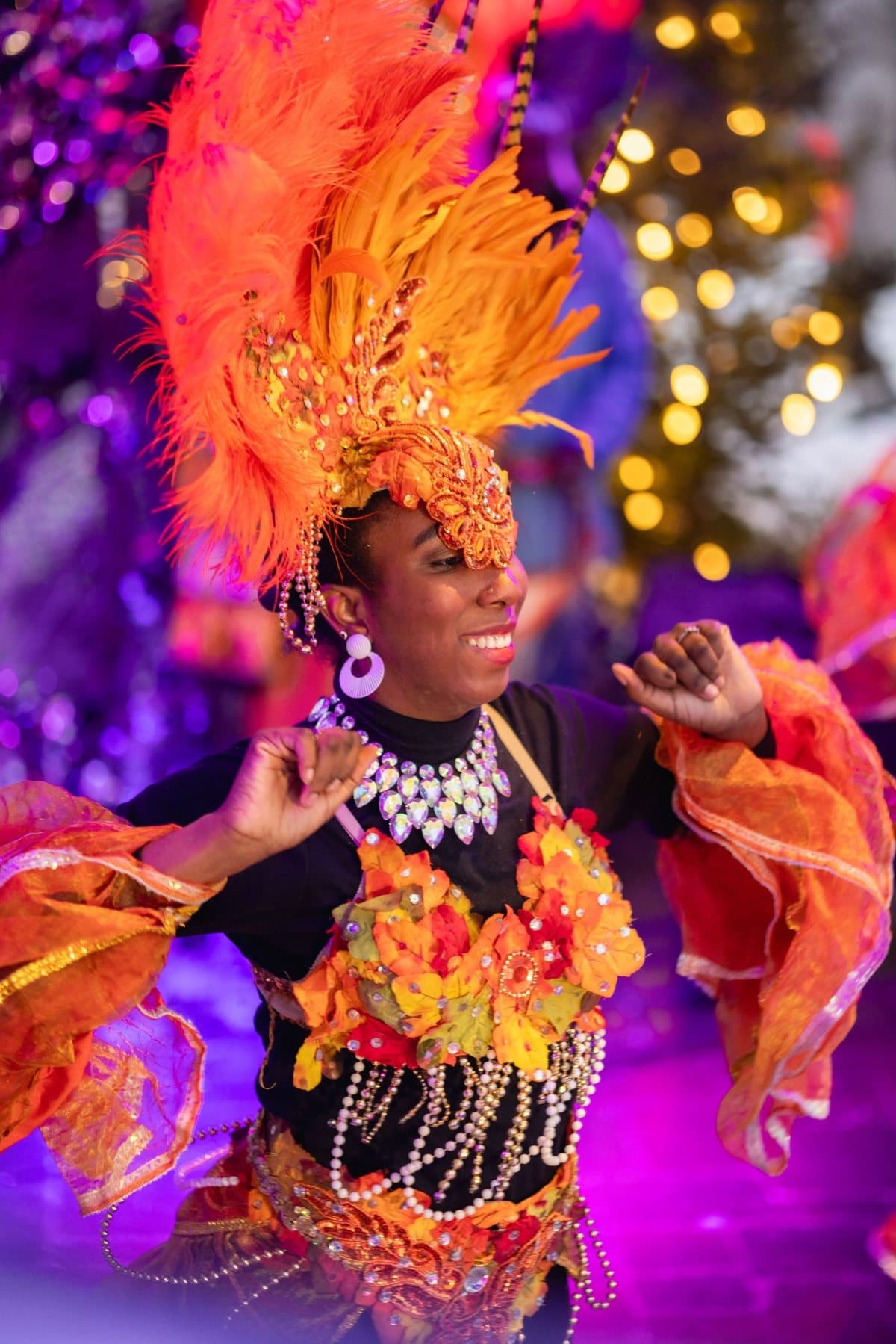 A carnival dancer in an orange feathered headdress and beaded costume, smiling, with festival lights behind