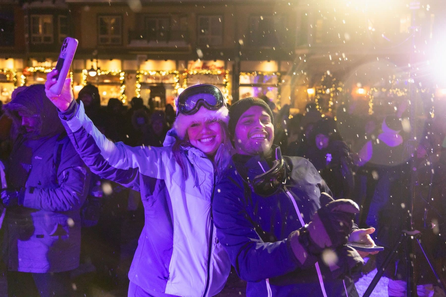 Two people in ski jackets taking a selfie in falling snow at night, the Village shopfronts strung with lights behind them