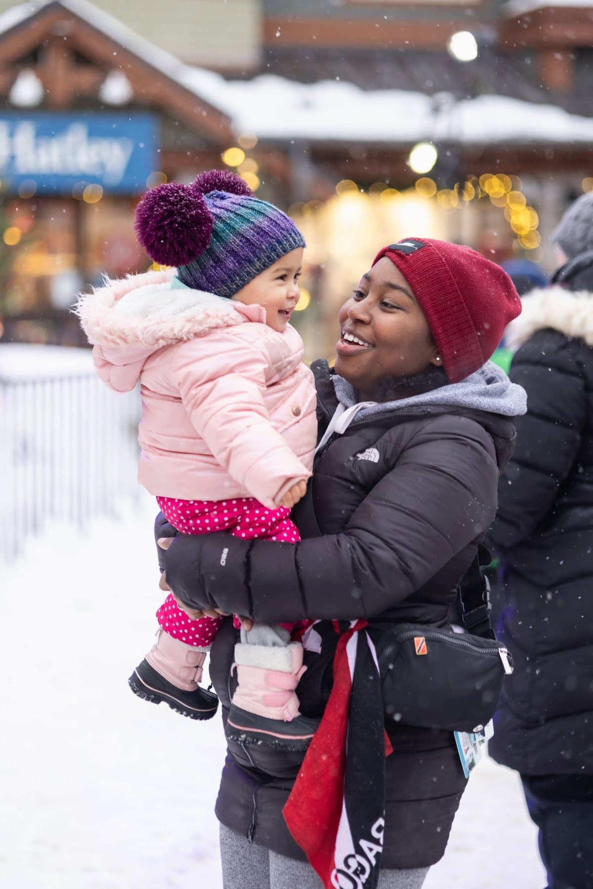 A woman in a red toque holding a laughing toddler in a pink snowsuit, snow on the Village rooftops behind