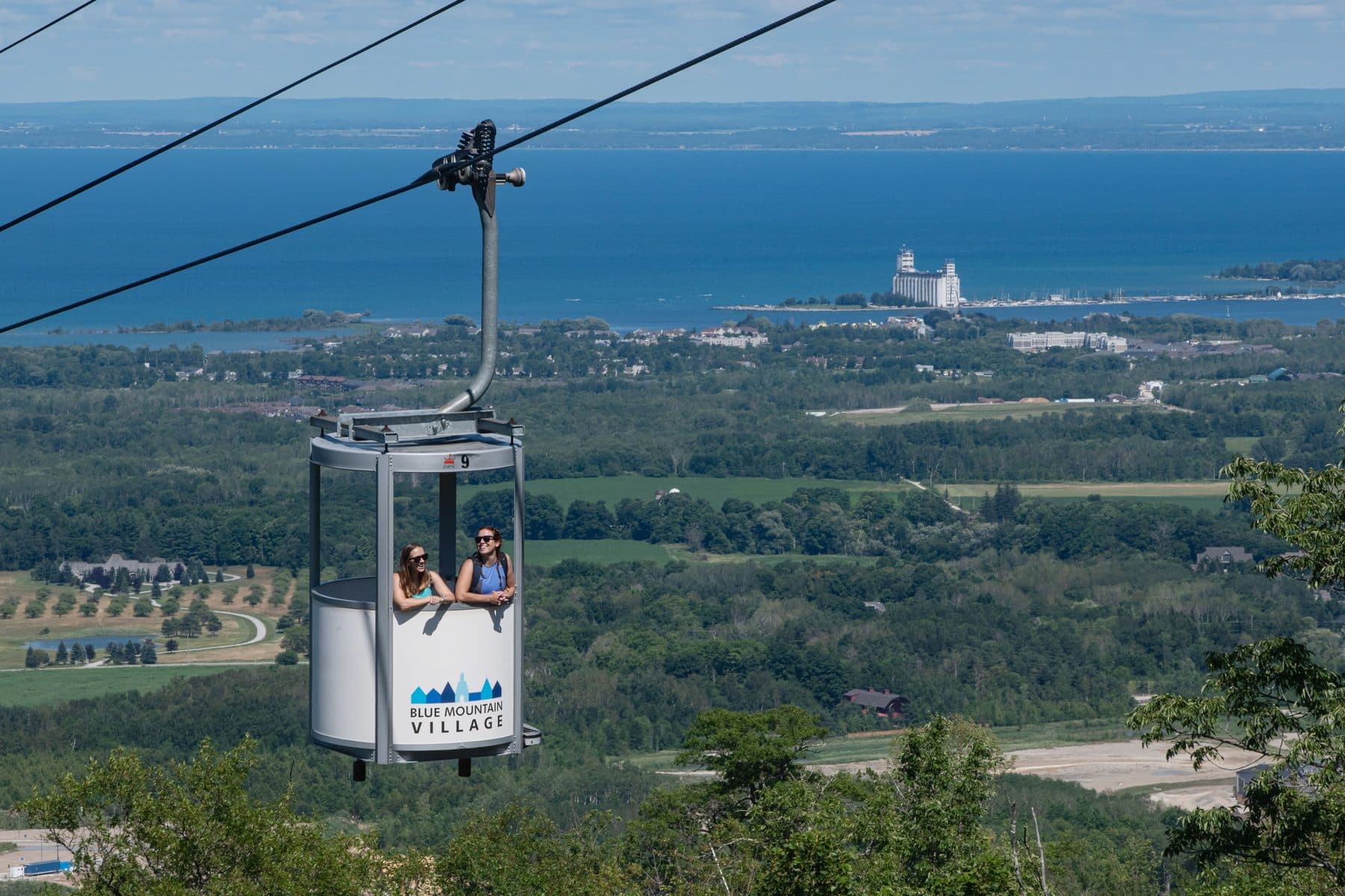 An open-air gondola cabin above the forested slope with Georgian Bay behind