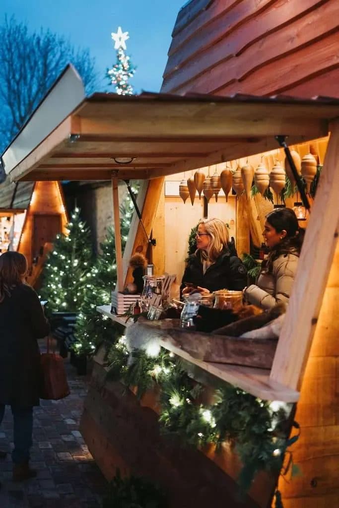 A lit cedar chalet at the Elora Christmas Market with two people inside serving customers at the counter