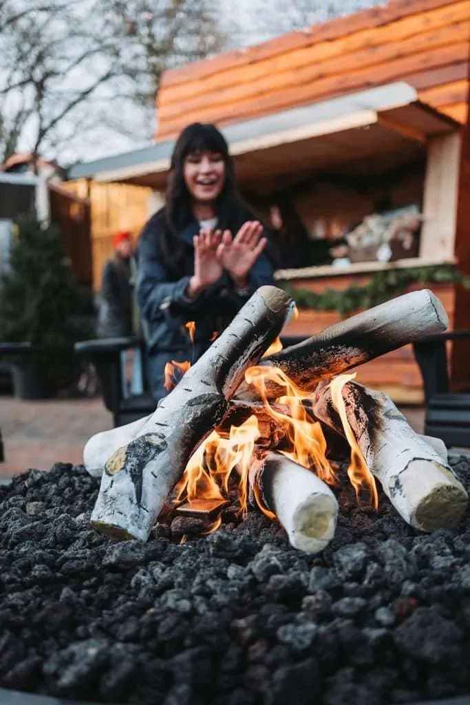 A woman in a winter coat warming her hands over an open fire table at the Elora Christmas Market after dark