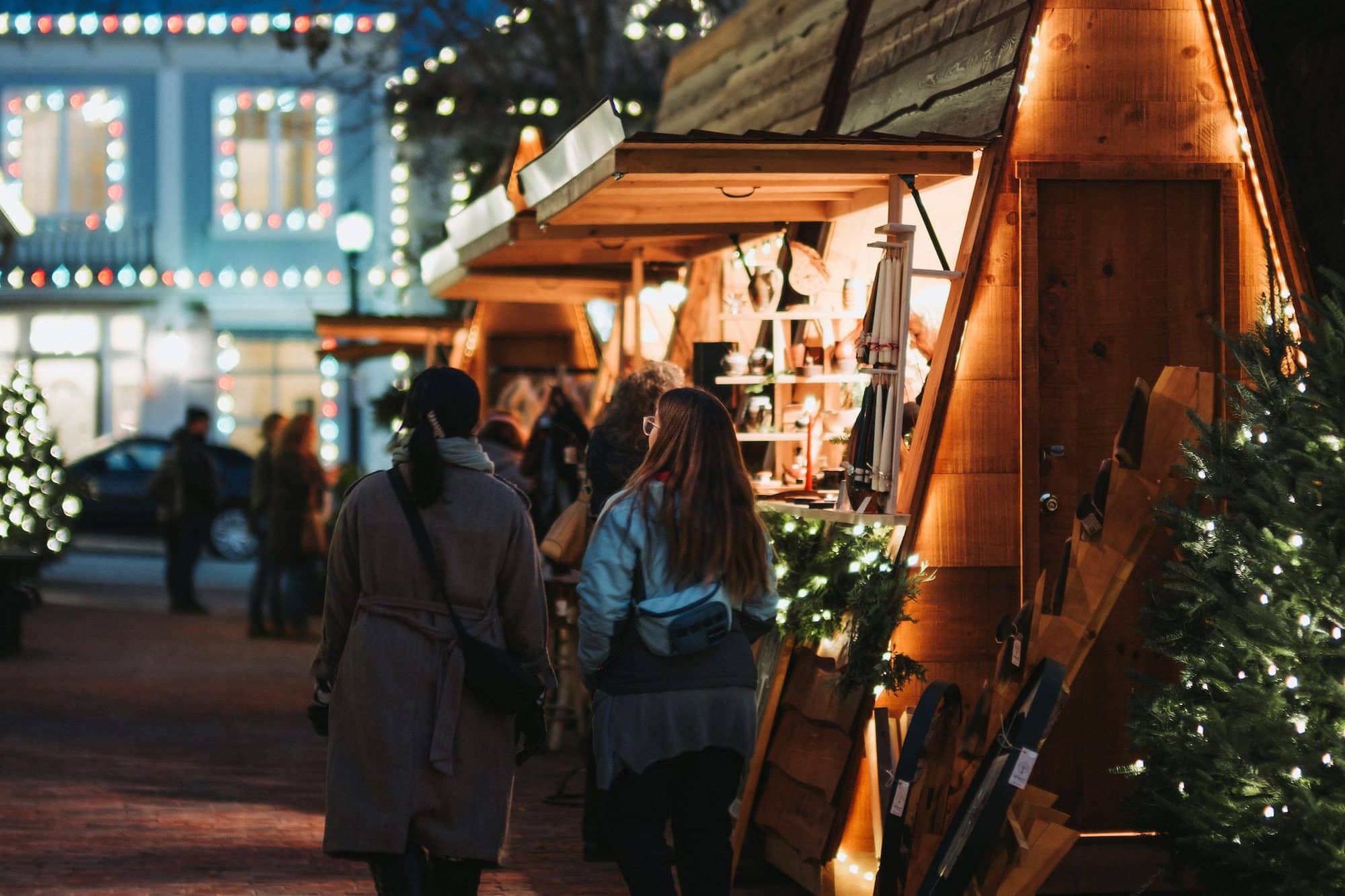 Shoppers walking between lit cedar chalets on an Elora street at dusk, strings of lights overhead