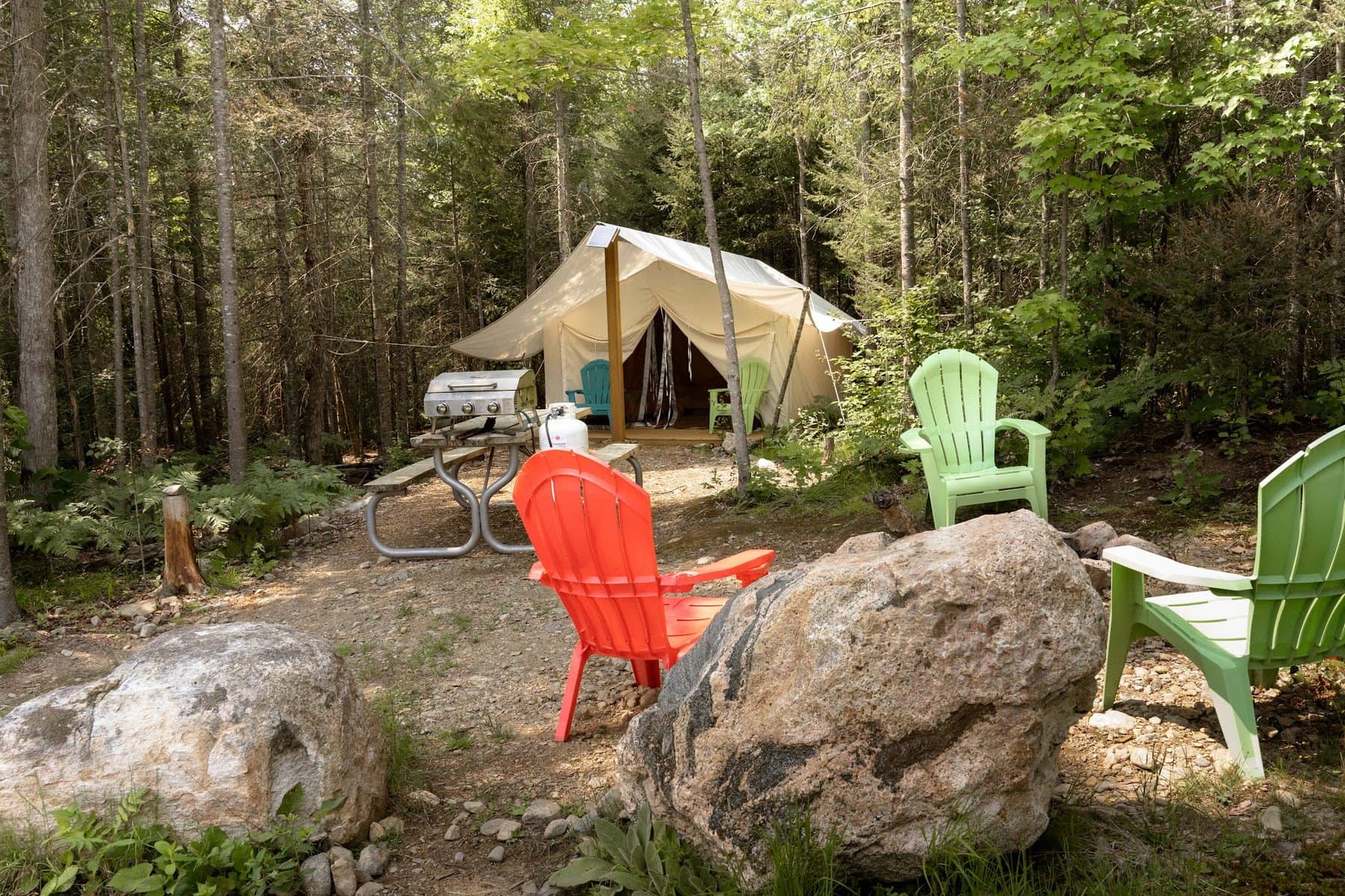 A site at Four Corners Algonquin: bright orange and green Muskoka chairs round a fire ring in the foreground, a picnic table and barbecue, and a canvas safari tent set back among the trees