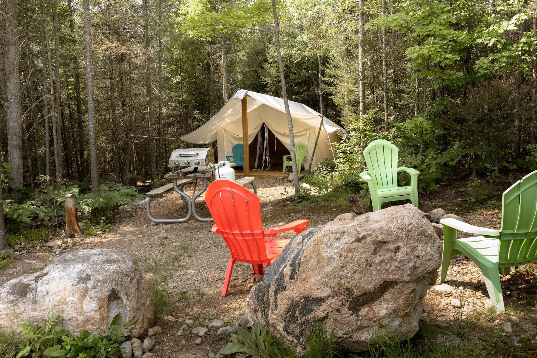 A site at Four Corners Algonquin: bright orange and green Muskoka chairs round a fire ring in the foreground, a picnic table and barbecue, and a canvas safari tent set back among the trees