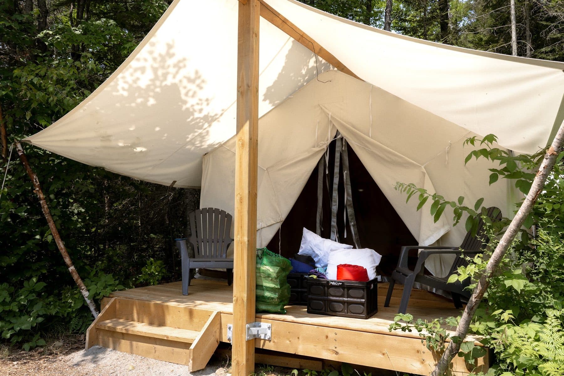 The open front of a safari tent on its raised deck at Four Corners Algonquin, with chairs either side and bedding stacked in a crate ready for arrival