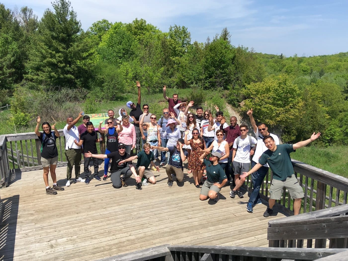 A large group on a wooden boardwalk lookout above a green valley, arms in the air