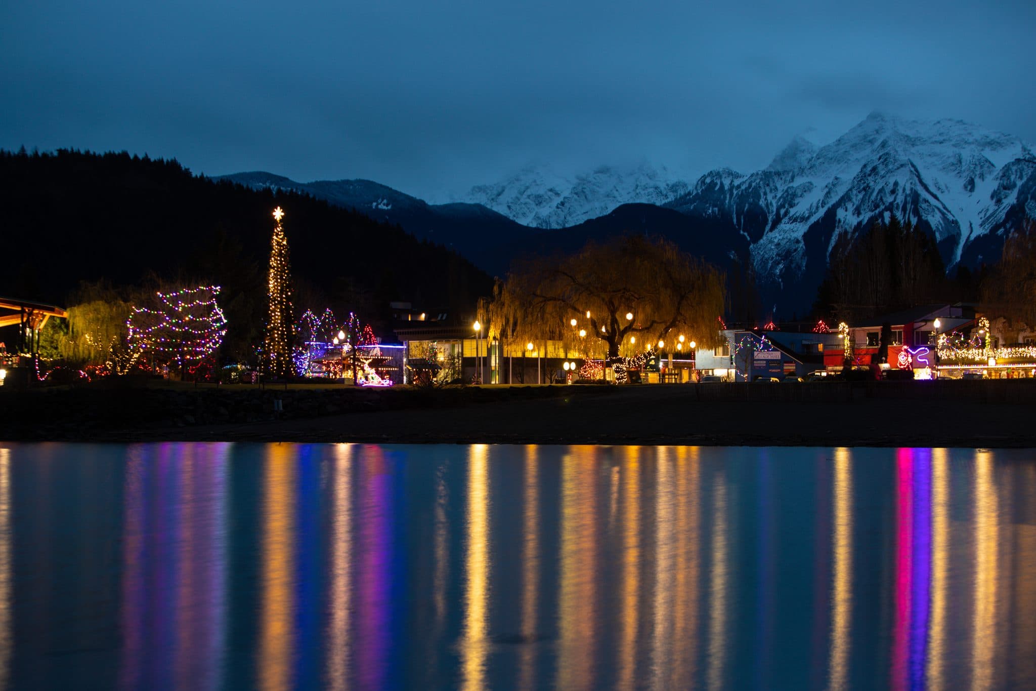 Harrison Hot Springs at night during Lights by the Lake, coloured lights along the shore reflected down the water with snowy mountains behind