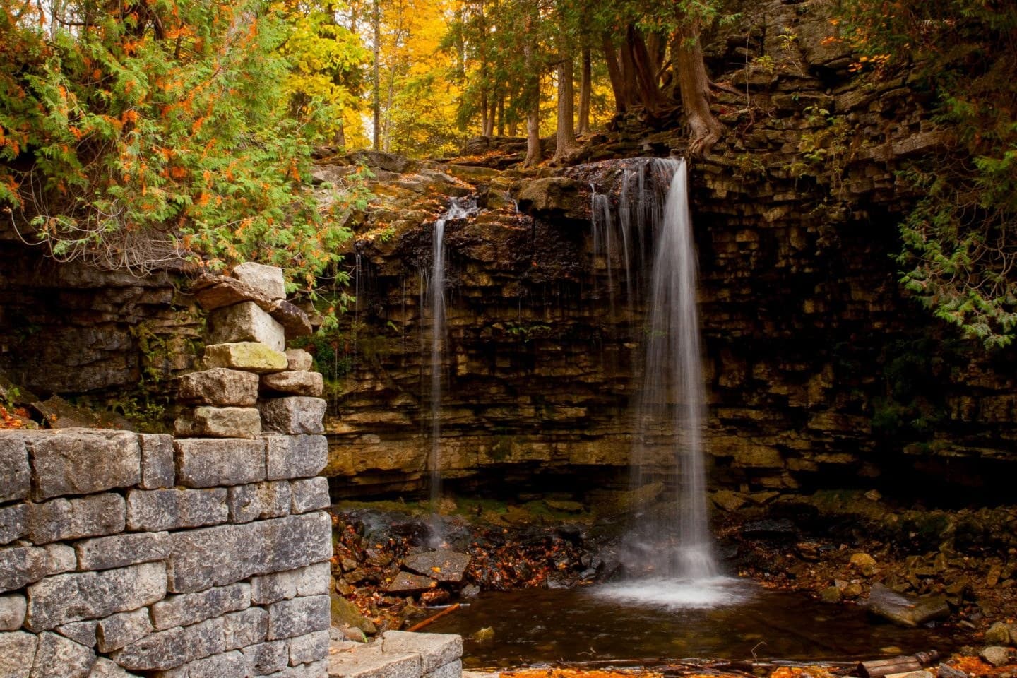 A waterfall dropping over a layered limestone ledge into a shallow pool, orange leaves on the rocks below and cedars and yellow maple above, with a dry stone mill wall in the foreground