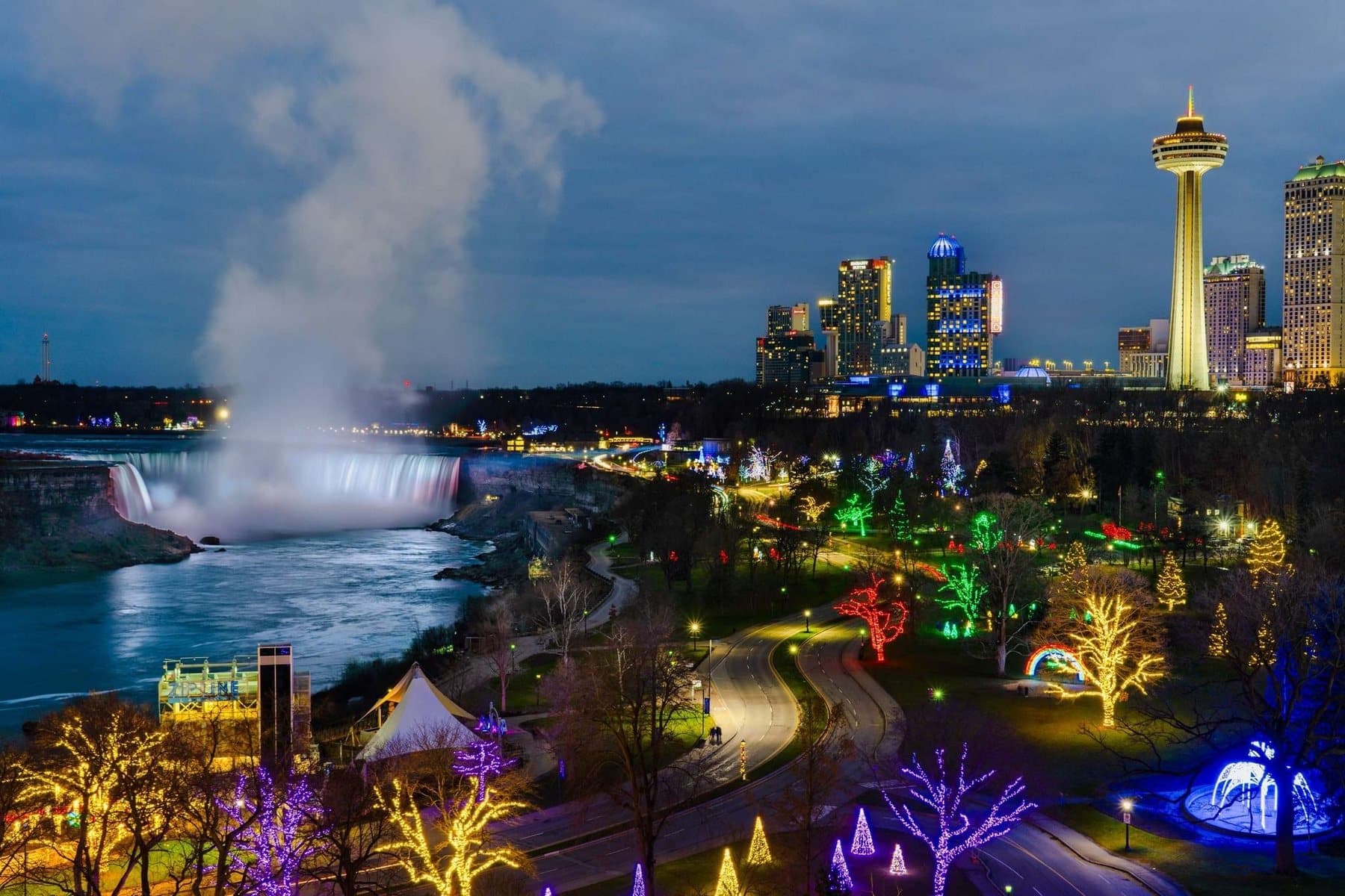 Niagara Falls at dusk from above, the Horseshoe Falls lit white and the parkland beside it strung with coloured lights, the Skylon Tower behind