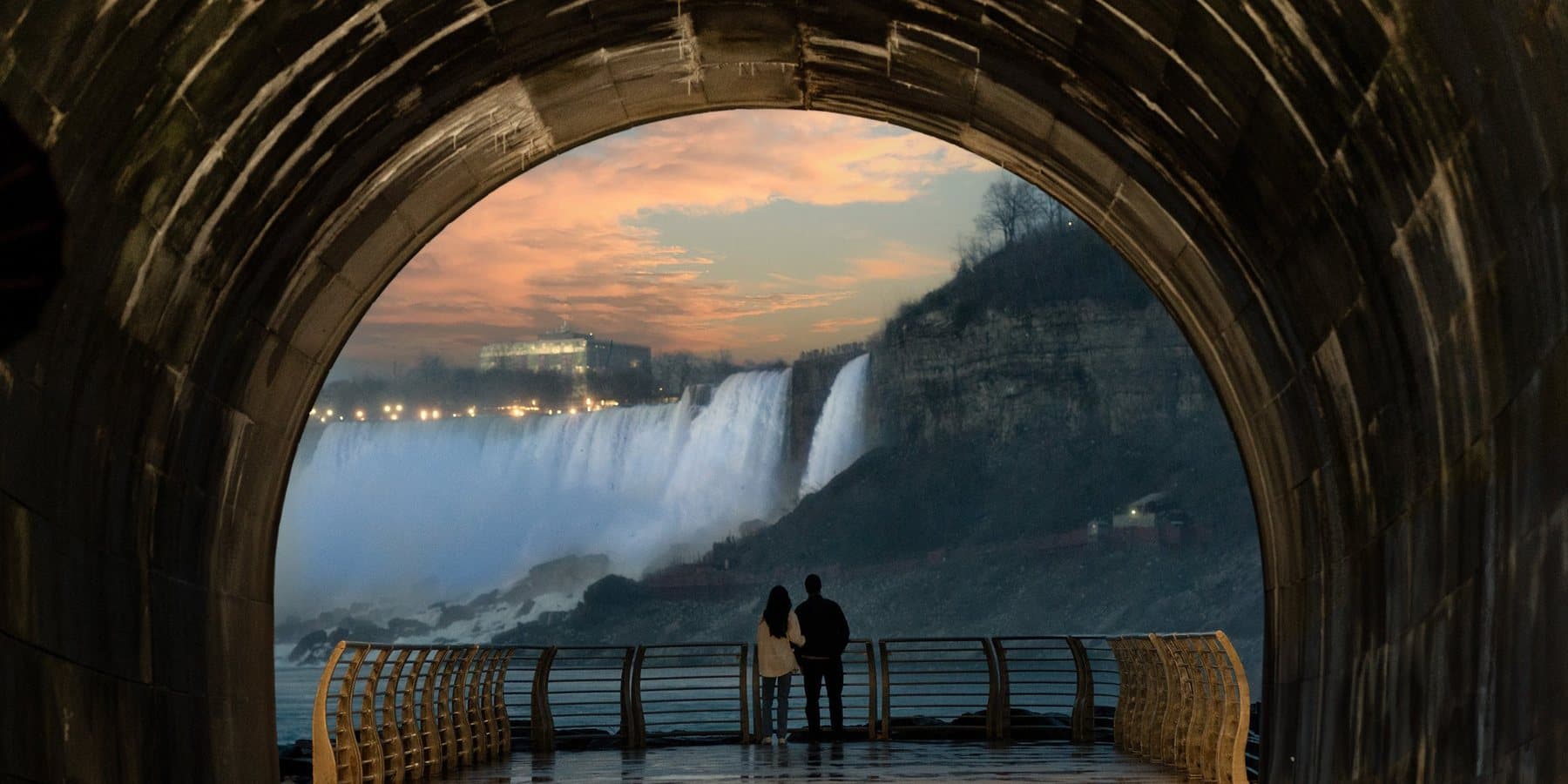 Two people on the deck at the end of the tunnel, looking out through the stone arch at the falls at sunset
