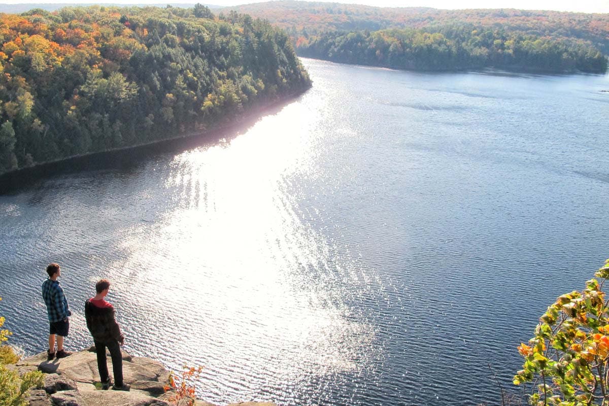 Two people standing on a rock ledge looking down a long lake in autumn colour