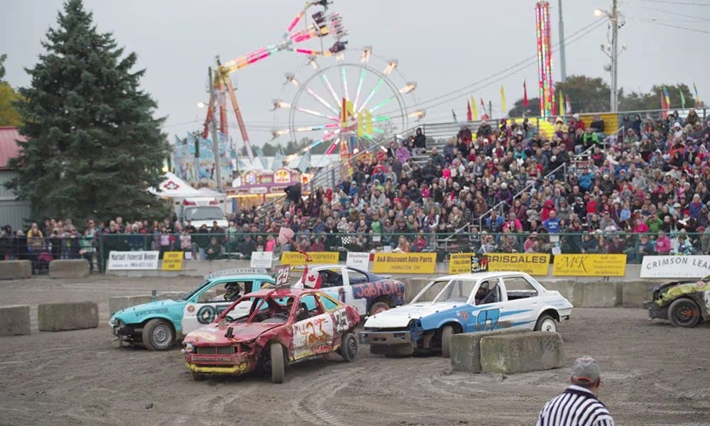 Four cars in the demolition derby ring at Rockton, a full grandstand behind them and the midway lit up beyond