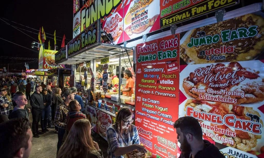 People queuing at a funnel cake and corn dog stand at night, the menu boards lit up