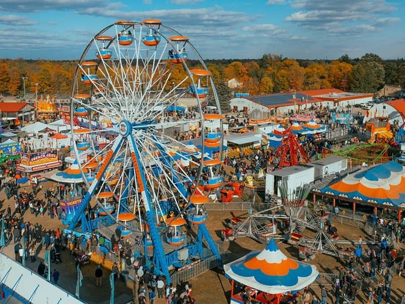 The Rockton fairground from above, the orange and blue Ferris wheel over rows of tents, with the trees behind it in full autumn colour