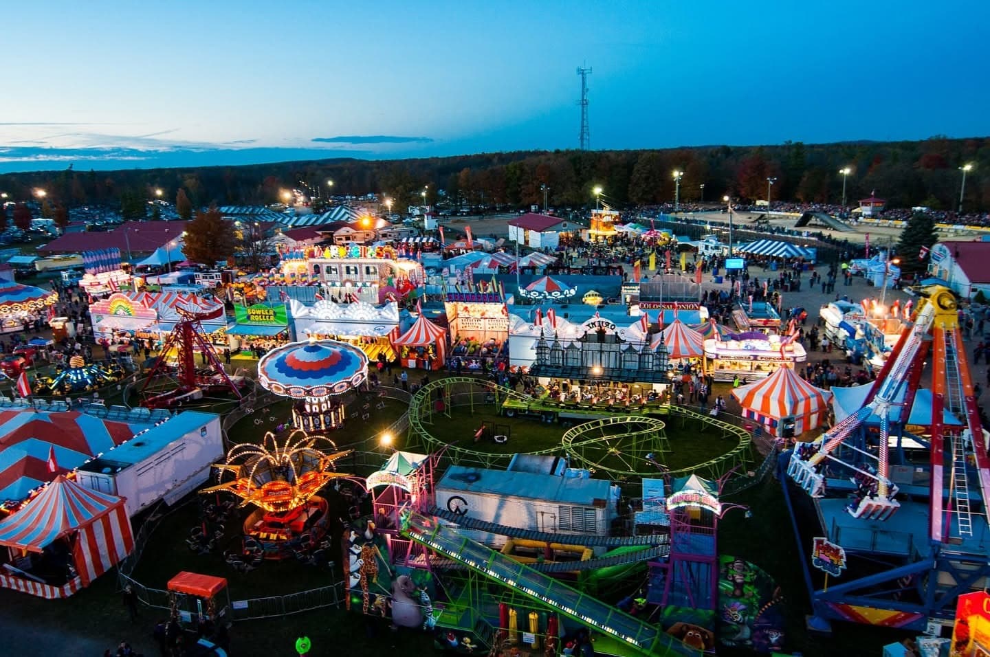 A fairground midway lit up at dusk, rides and striped tents packed with people, autumn trees along the far side