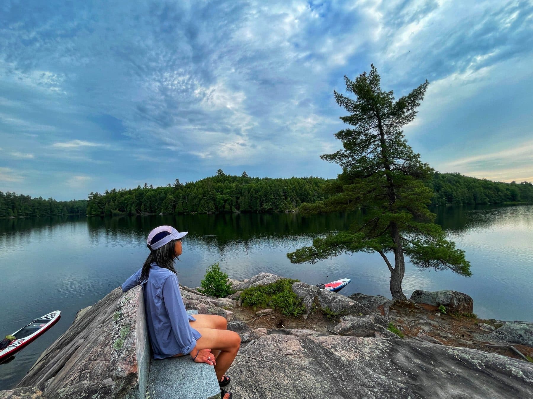 A woman in a sun hat sitting on granite at the water's edge, a paddleboard beached beside her