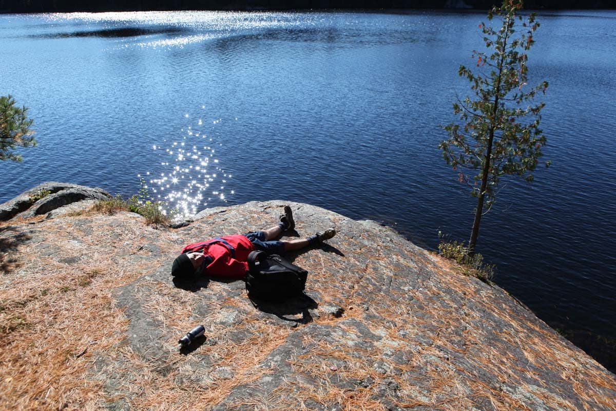 A person lying flat on a slab of granite beside sunlit water, arms out