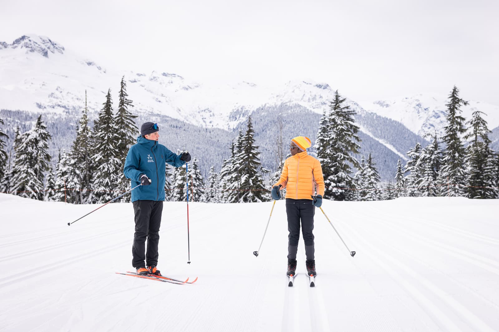An instructor and a learner side by side on a groomed track with mountains behind