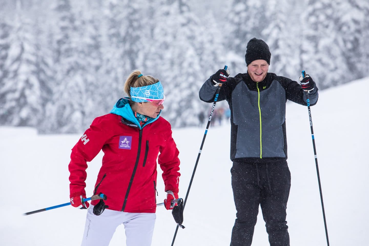 An instructor in a red jacket skiing beside a learner through snowy trees