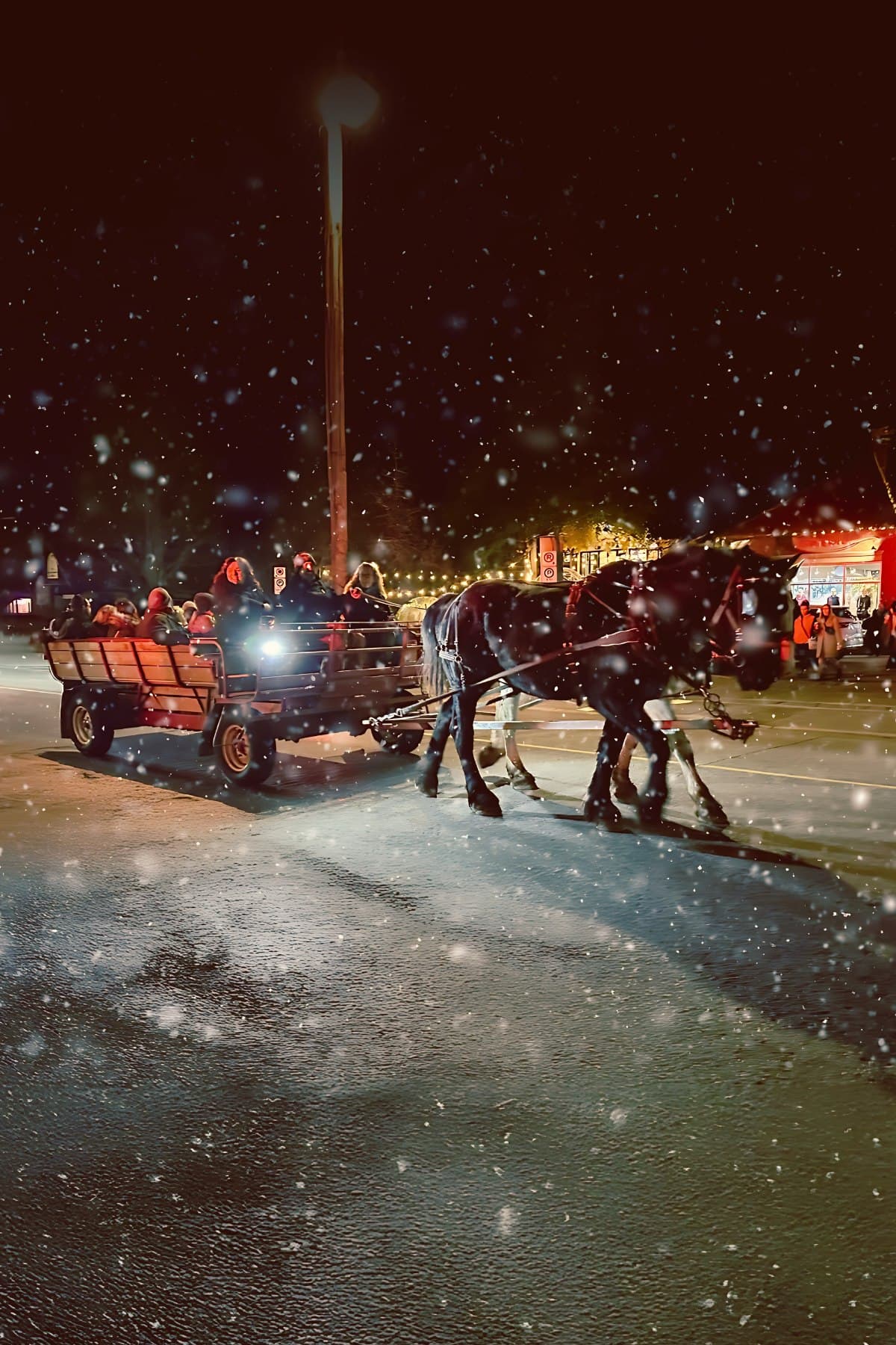 A horse drawn trolley full of people moving through falling snow on a lit village street at night
