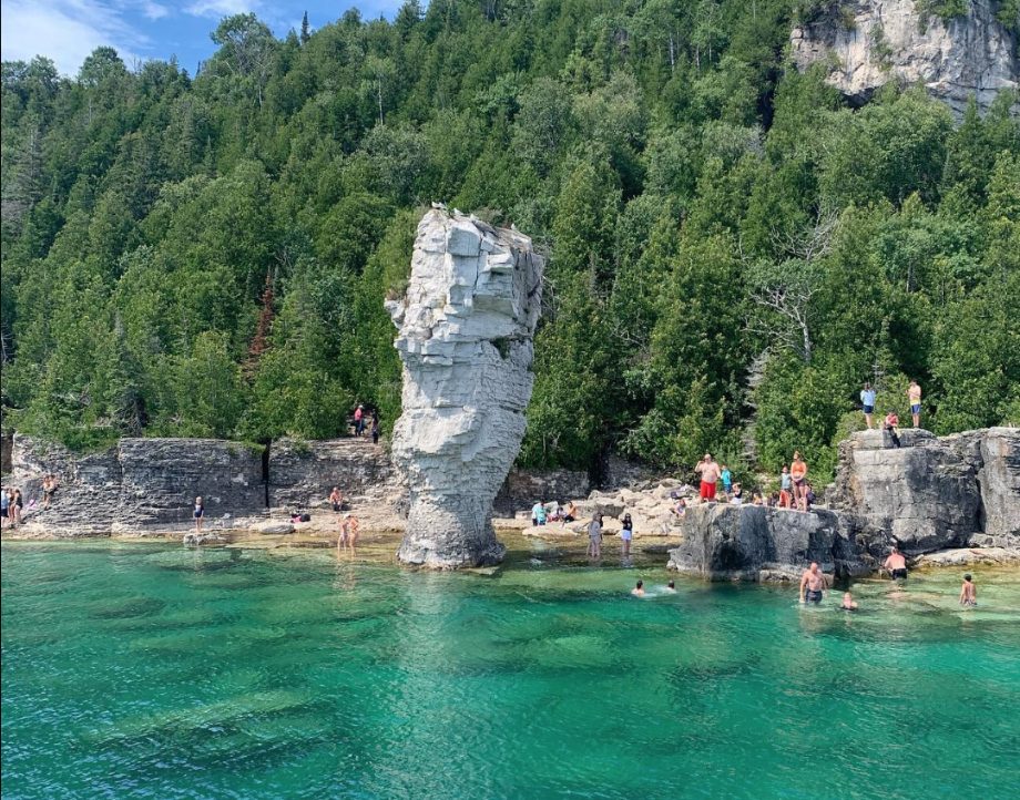 A limestone rock pillar rising out of turquoise water, with people swimming and sitting on the rocks