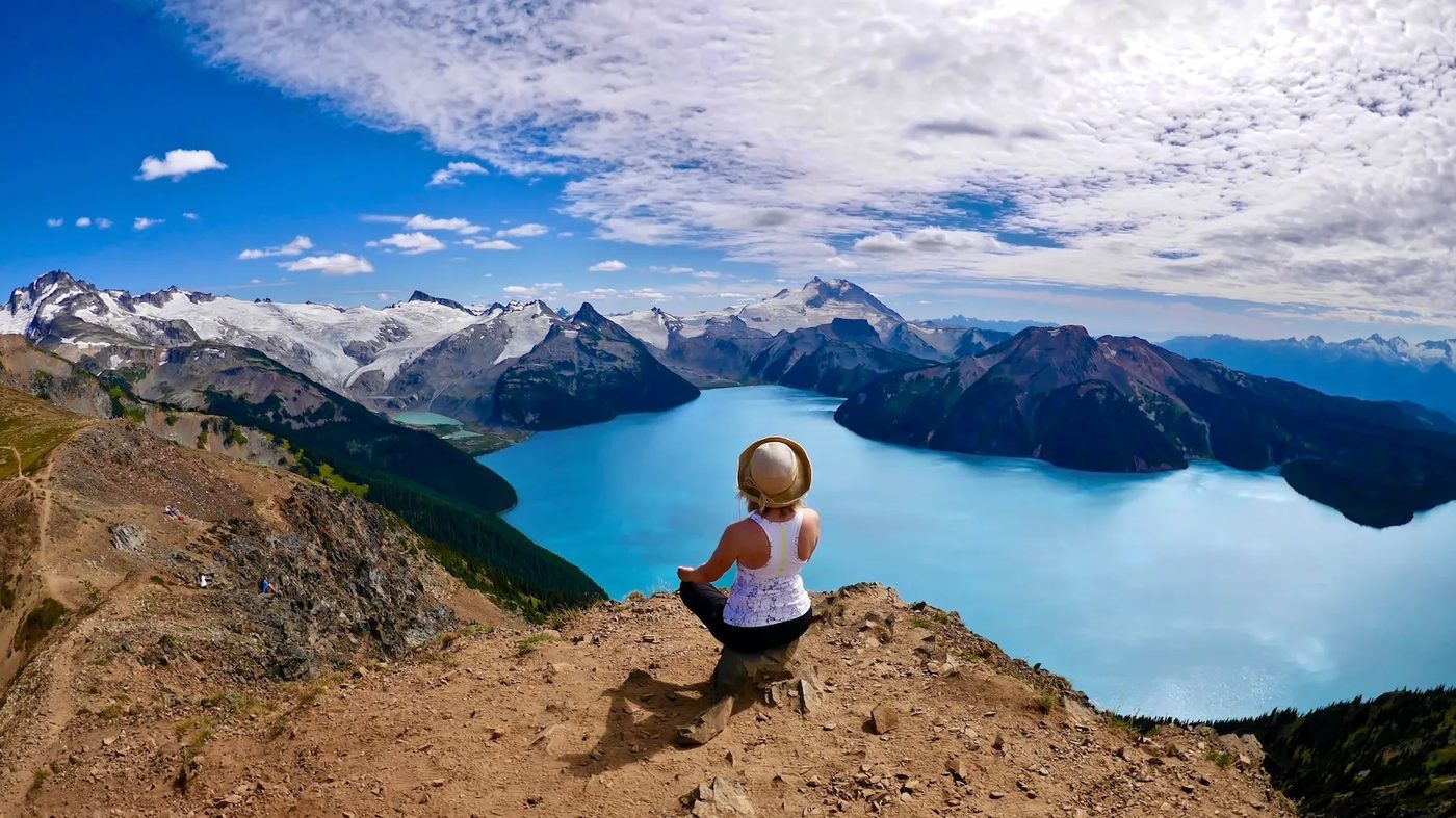 A hiker sitting on a rocky ridge, looking down at a turquoise mountain lake