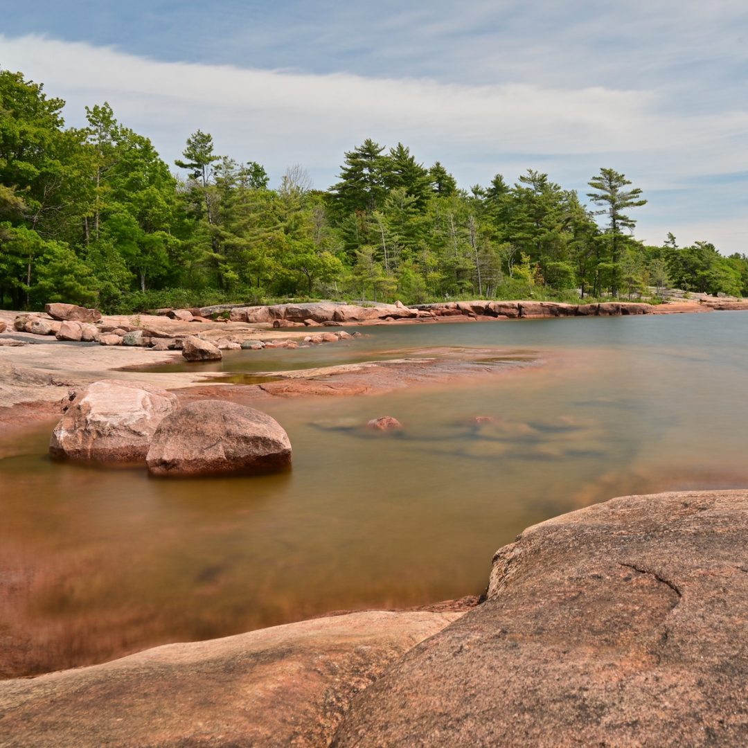 Pink granite rocks and shallow clear water along a wooded shoreline