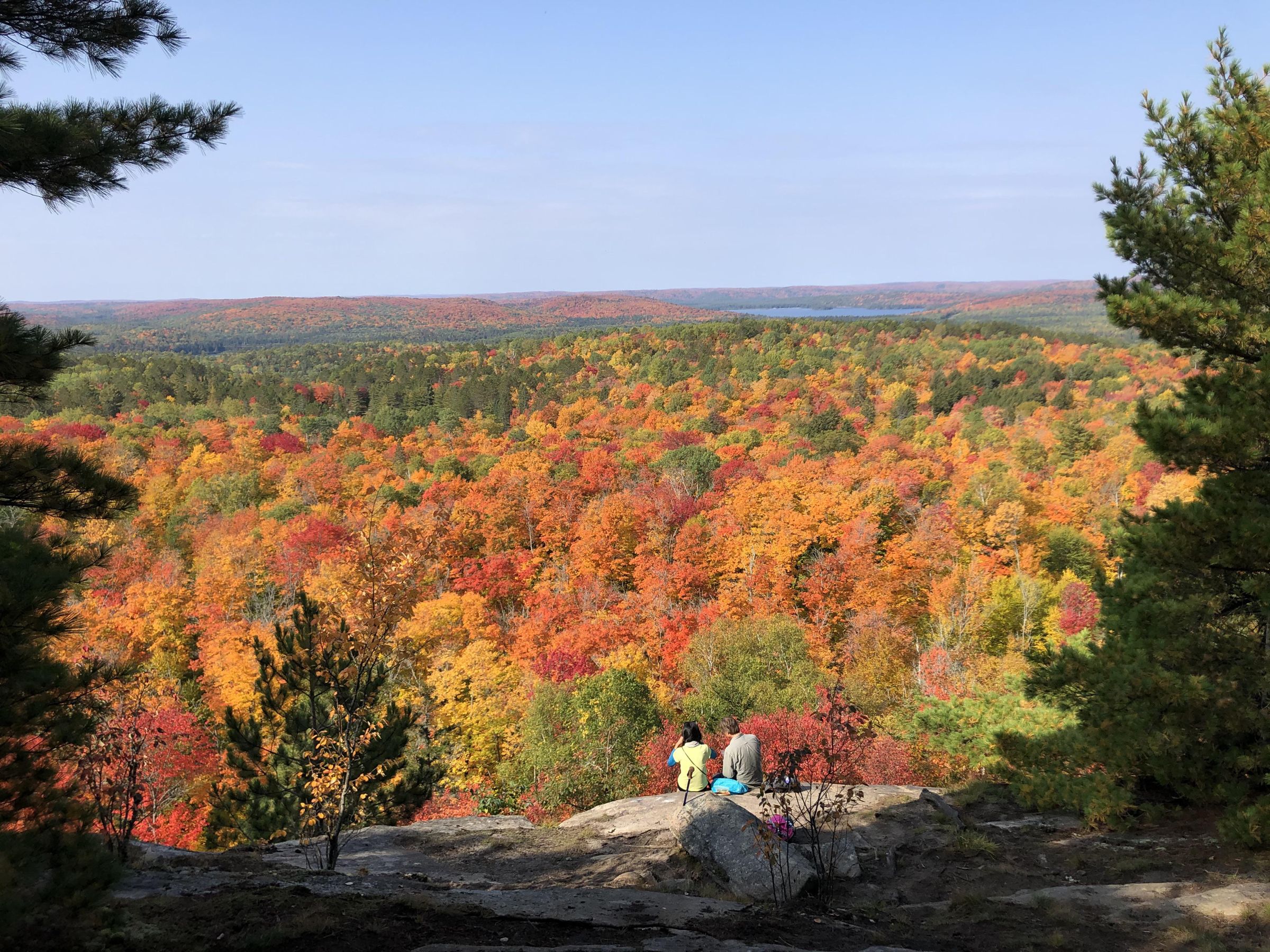 Two hikers sitting on a rock lookout above a valley of red, orange and yellow fall forest