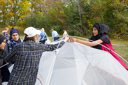 Four women, two of them in headscarves, raising the poles of a dome tent together in a grassy clearing with autumn trees behind them