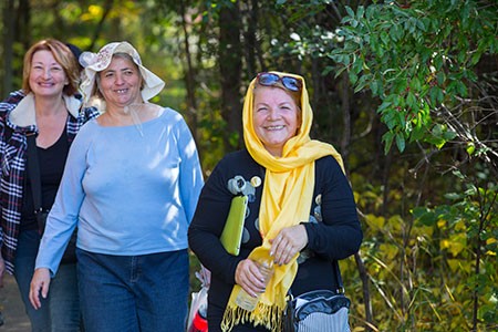 Three women walking a woodland trail in autumn, laughing, the one in front wearing a bright yellow headscarf